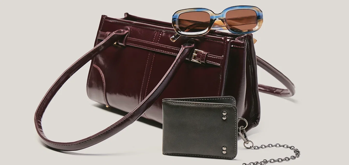 Bolso de mano de cuero burdeos oscuro con gafas de sol encima y una cartera negra con cadena y llave adjunta al frente.