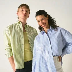 Dos personas están de pie sonriendo frente a un fondo liso, vistiendo camisas de rayas holgadas y pantalones casuales, posando con confianza.