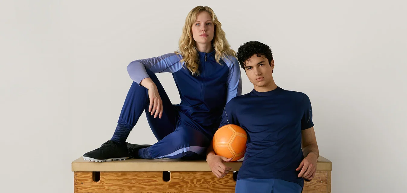 Dos personas con ropa deportiva azul posan con una pelota naranja sobre una caja de gimnasia de madera contra un fondo claro y liso.
