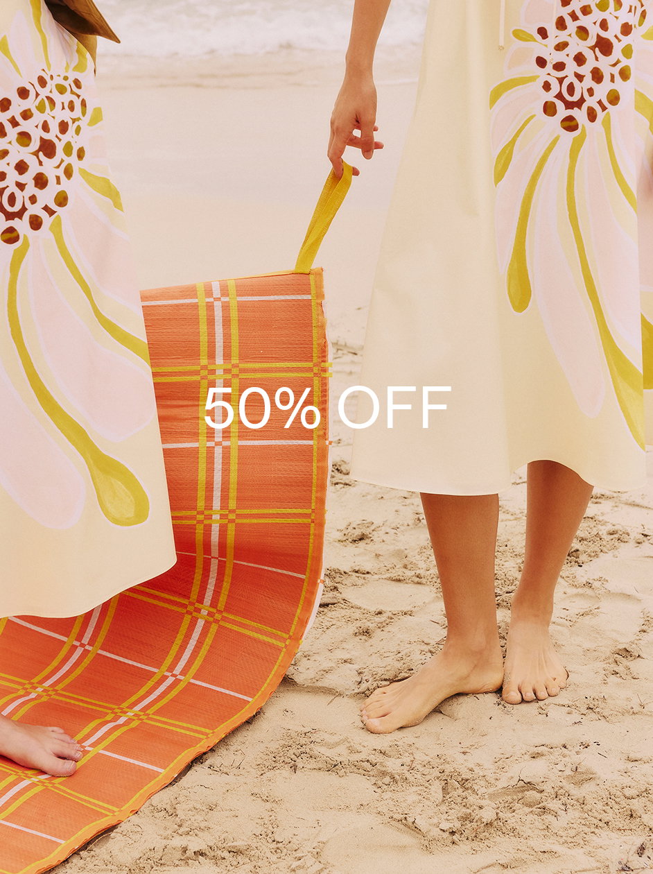 Ein Sale-Motiv von DOROTHEE SCHUMACHER mit dem zentrierten weißen Schriftzug '50% OFF'. Im Hintergrund stehen zwei barfüßige Models am Sandstrand in hellen Kleidern mit einem großen, abstrakten Sonnen-Print, die eine orange-gelb karierte Strandmatte halten.
