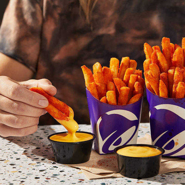 A hand holding a few Taco Bell Nacho Fries, dipping them into Nacho-Cheese Sauce, with 2 containers of Nacho Fries on the table.