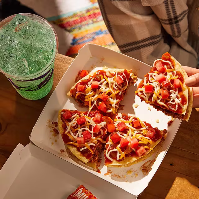 A Taco Bell Mexican Pizza and beverage on a table, with a hand reaching for a slice of Mexican Pizza.