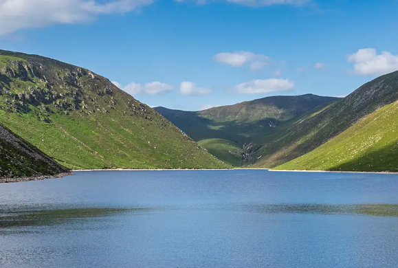 A serene lake surrounded by green hills under a blue sky with scattered clouds.