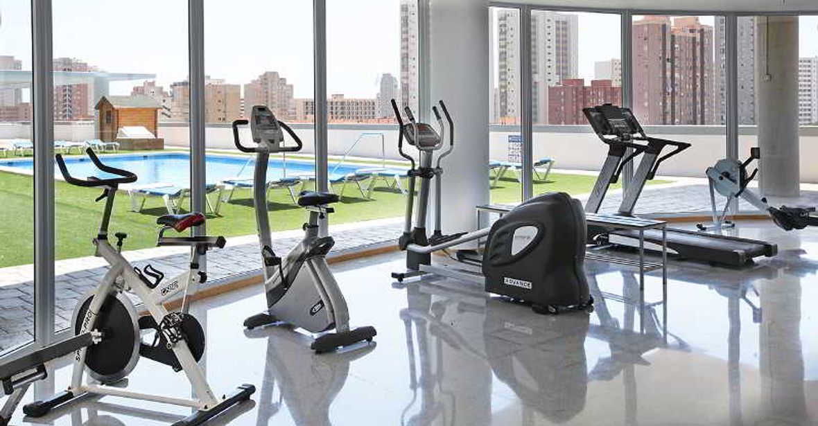 Bright gym with exercise bikes, cross-trainers, treadmill and rowing machine by floor-to-ceiling windows overlooking rooftop pool and city skyline