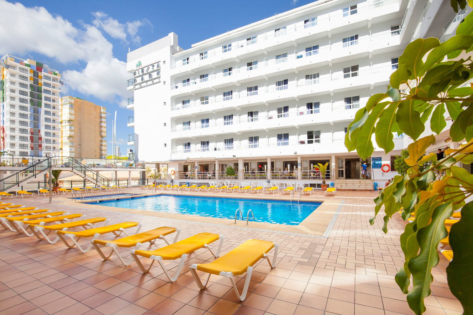 Outdoor hotel swimming pool with rows of yellow sun loungers on a tiled terrace in front of a modern white multi-storey hotel under a blue sky
