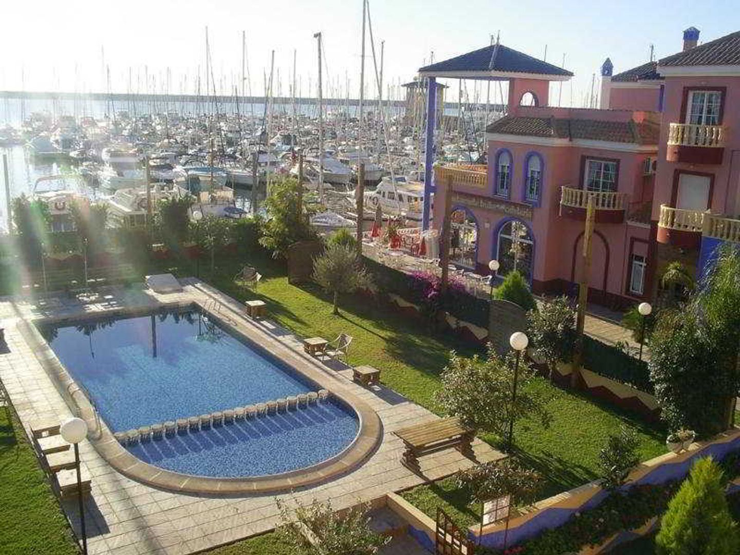 Sunlit marina with dozens of moored sailboats, foreground outdoor swimming pool and pink Mediterranean-style hotel building