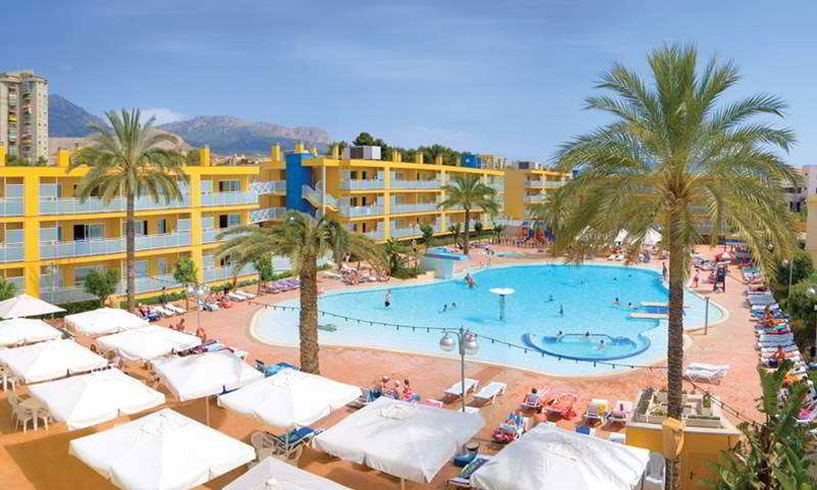 Resort hotel with large outdoor swimming pool, palm trees, white sunbeds and umbrellas, yellow apartment building and distant mountains