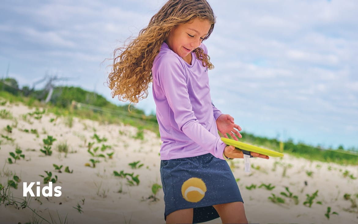 A little girl wearing colorful clothes on the beach, playing with a ball and racket with text: Kids