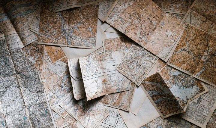 Several old, overlapping paper maps are spread out, showing various geographic locations, marked borders, rivers, and cities, with a sepia-toned color palette.