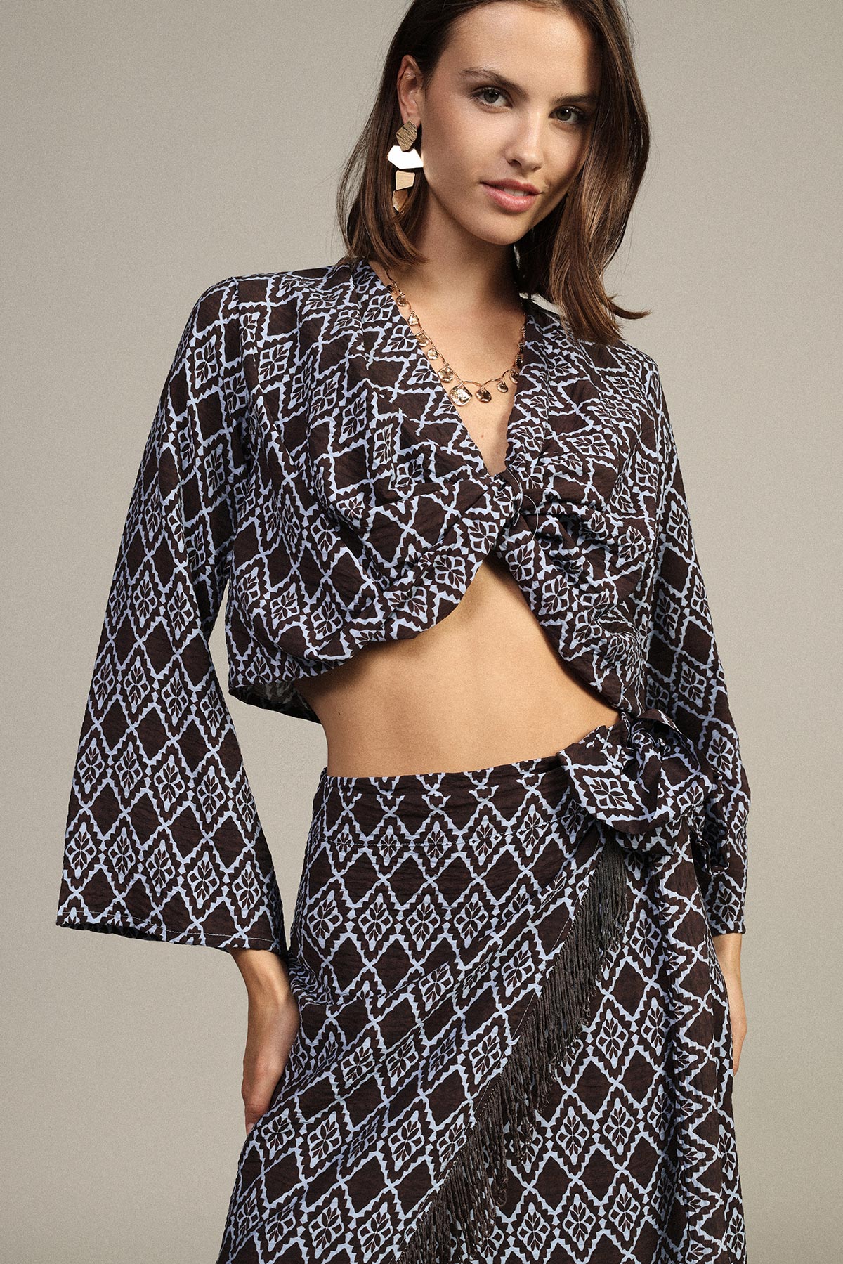 Woman model wearing black and light-blue geometric patterned wrap crop top and matching skirt with fringe, gold jewellery, neutral background