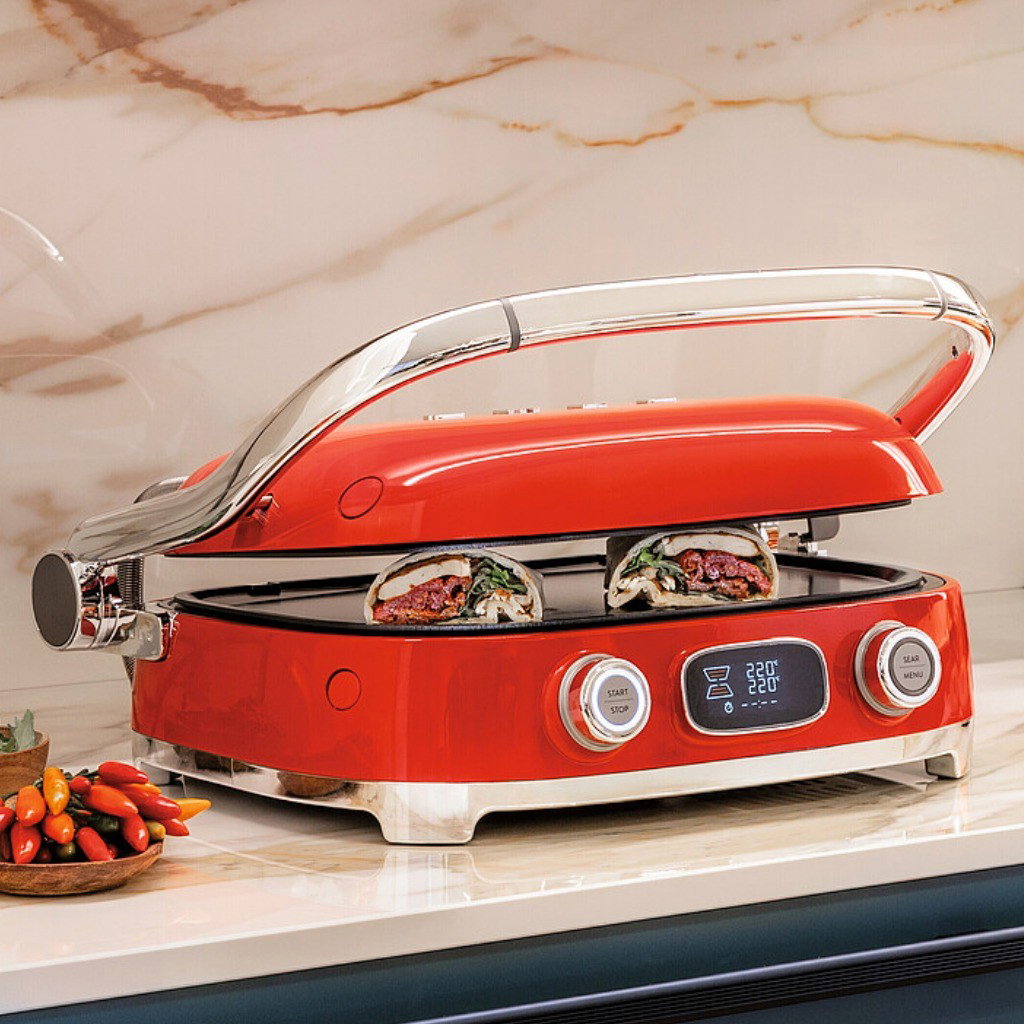 Grill Multiusos Smeg rojo cocinando brochetas y verduras con la tapa parcialmente cerrada sobre una encimera de cocina
