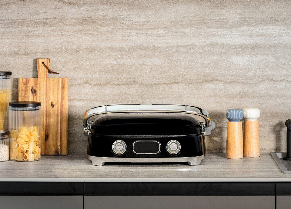 Grill Multiusos Smeg negro sobre una encimera de cocina, rodeado de accesorios de madera y tarros de almacenaje