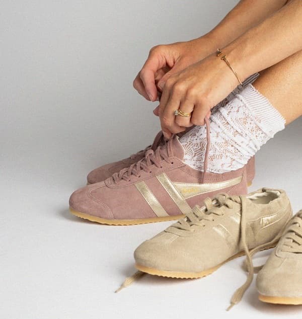 An image of a woman wearing a pair of Gola Aurora in brownrose & next to her feet there's also a pair in bone/gold