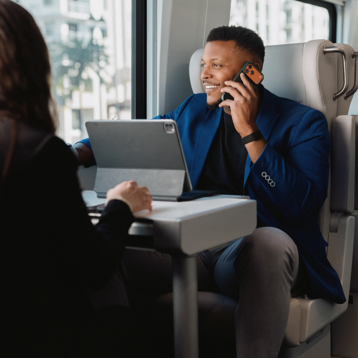 A man in a blue blazer talks on a smartphone while seated at a table on a modern train.