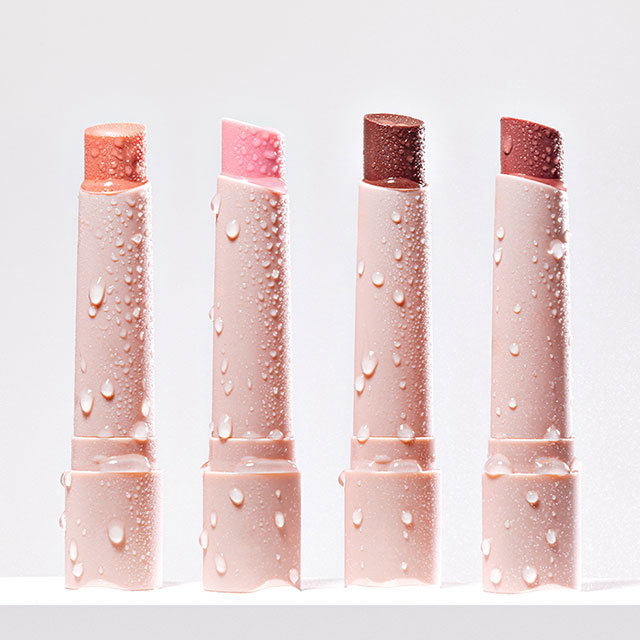 Four pink lipsticks with water droplets, arranged upright on a white surface.