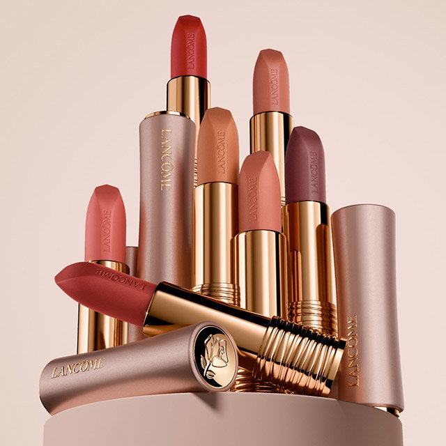 A collection of Lancôme lipsticks standing upright on a circular surface. The lipsticks feature various shades, including red, pink, peach, and mauve, with gold and rose gold casing. One lipstick is horizontally placed with its cap off, showcasing the embossed Lancôme logo on the base. The background is a soft, muted pink.