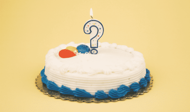 A white frosted cake with blue icing accents and a question mark candle on top against a yellow background.