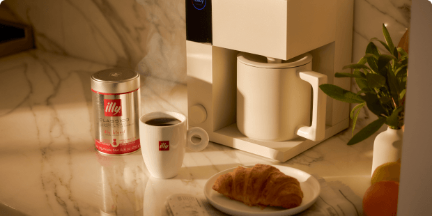 illy coffee, a freshly brewed cup, and a croissant arranged beside a coffee machine on a marble countertop