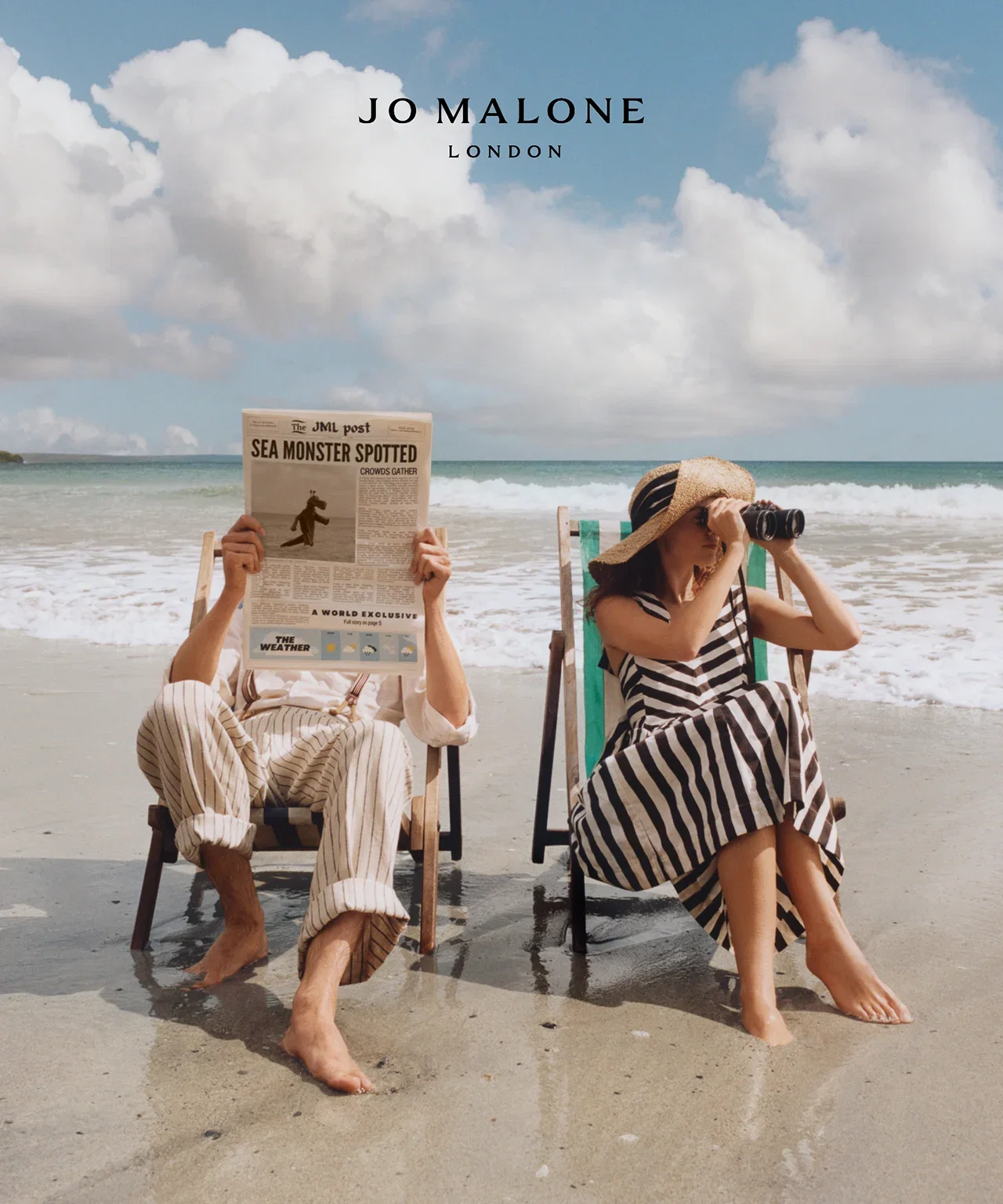 Two people in striped clothing sit on beach chairs by the shore; one reads a newspaper, the other looks through binoculars. Waves gently roll in the background.