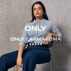 Femme aux longs cheveux assise sur une chaise, portant un pull bleu clair, un pantalon bleu foncé et des mocassins bruns, devant un mur industriel gris.