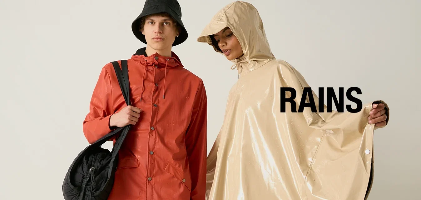 Deux personnes portant des vêtements de pluie : l'une en veste rouge et chapeau seau noir avec un sac, l'autre en poncho beige à capuche et shorts noirs.