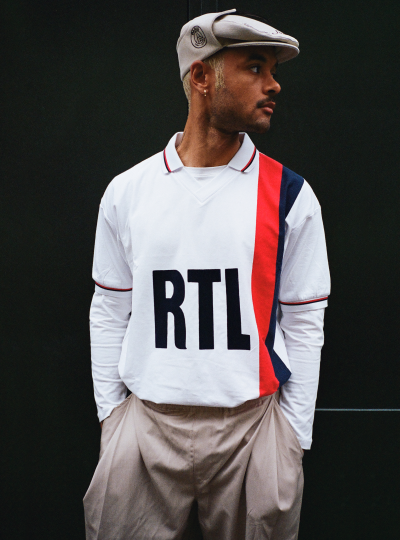 Model wearing a retro-inspired PSG RTL shirt with red and navy stripe detailing, styled with a flat cap and neutral trousers.