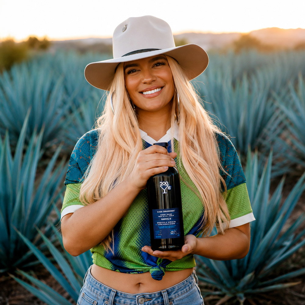 Smiling woman in a wide-brimmed hat holding a dark tequila bottle in an agave field at sunset