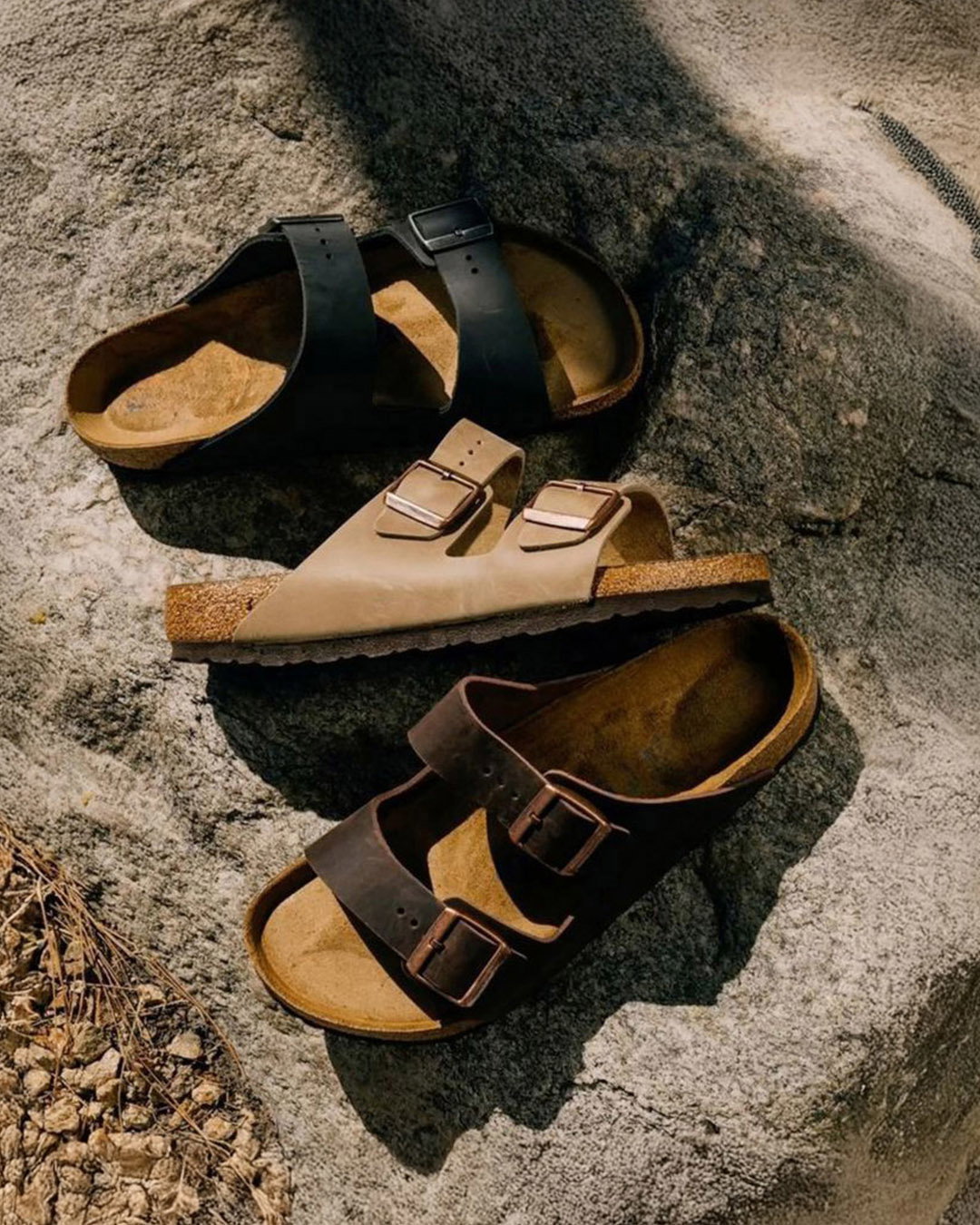 Three leather double‑strap buckle sandals in black, beige and brown arranged on a sunlit textured rock