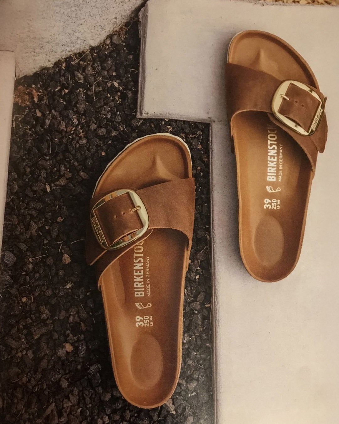 Pair of brown Birkenstock sandals with large gold buckles (size 39) resting on gravel beside a concrete slab