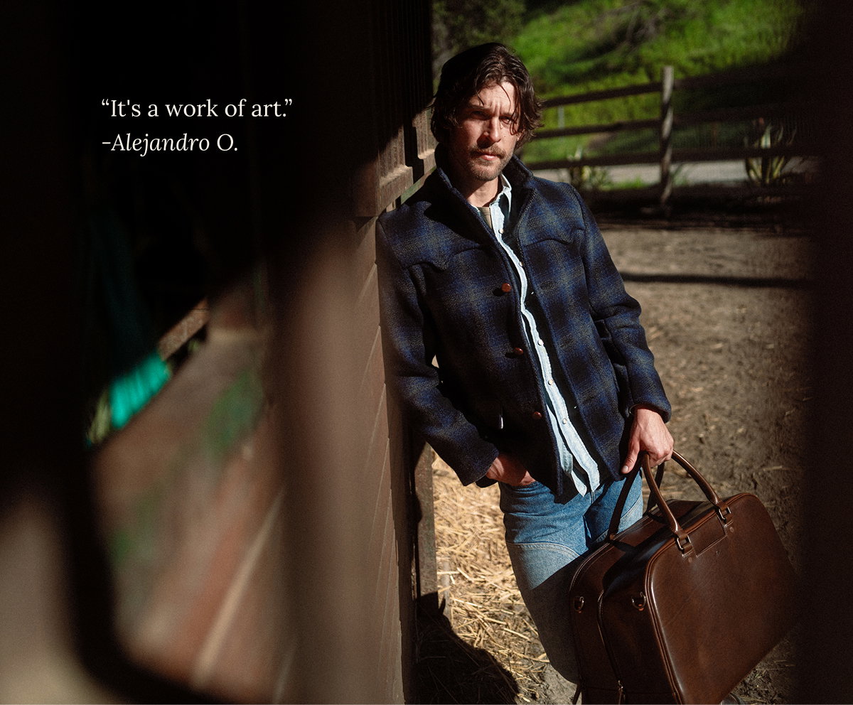 Man leaning against a barn wall holding a Satchel & Page leather Weekender — "It's a work of art." - Alejandro O.