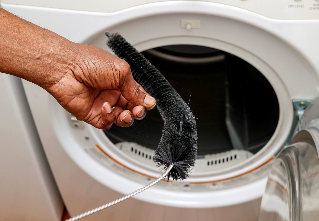 Hand holds a vent cleaning brush in front of an open dryer