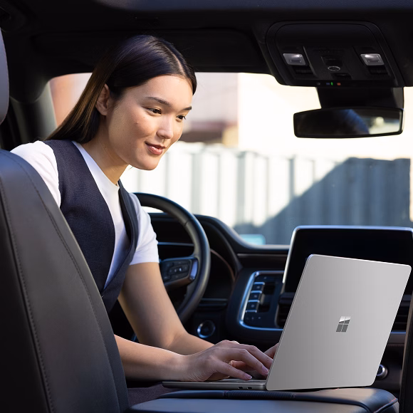 Shows a woman sitting in a car, working on her Surface Laptop which is propped on the center console.