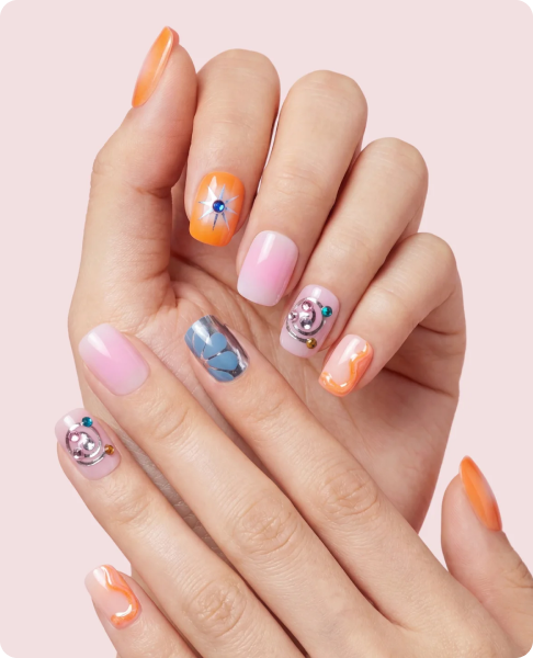 Close-up of two hands with mixed-pattern manicured nails in orange, pink, and blue, featuring marble effects, jeweled accents, and an orange sunburst design on the accent nails.