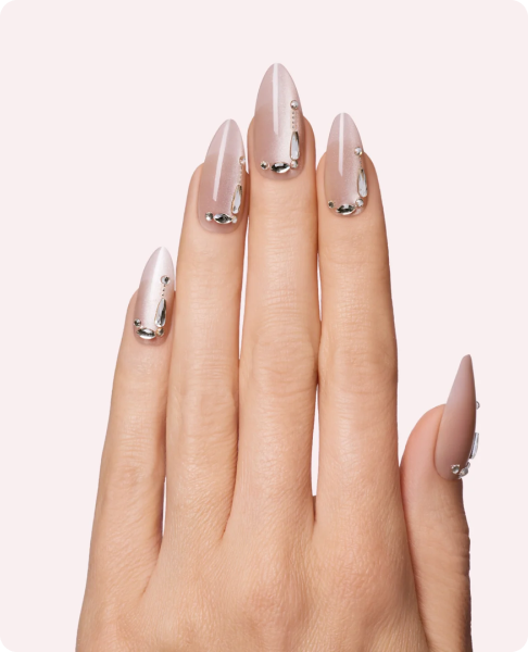 Close-up of a hand with long, almond-shaped manicured nails in a nude-to-mauve ombré, accented with silver metallic foil detailing and rhinestones near the cuticles.