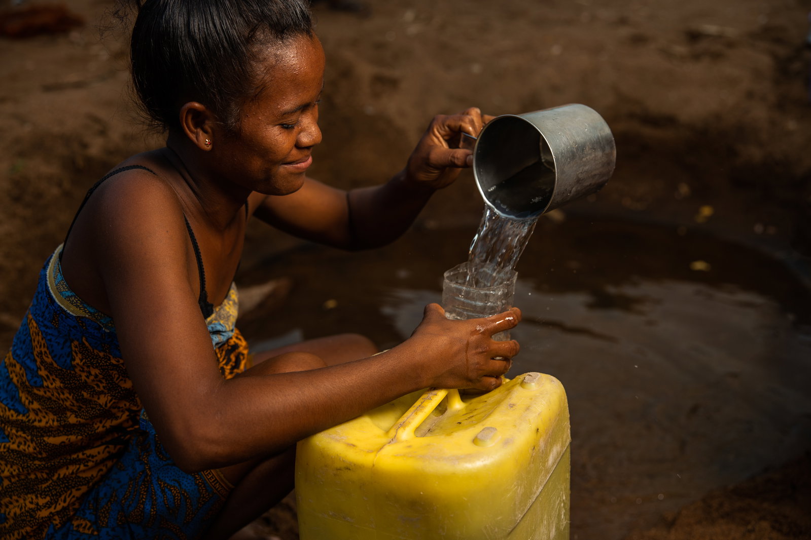 A woman pouring water into a glass that she got from a yellow jerry can.