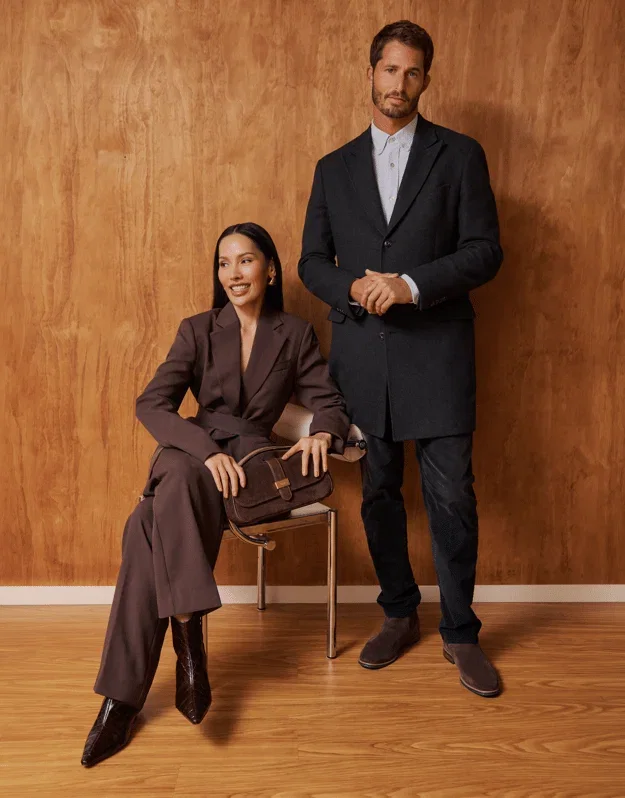 A medium shot shows a woman seated on a chair and a man standing, both wearing business attire against a wood-paneled wall. The woman wears a brown su...