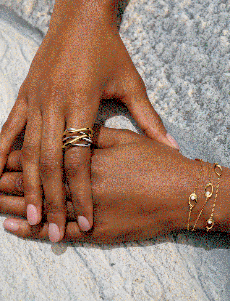 A pair of hands overlap and display an intertwined ring and pearl bracelet.