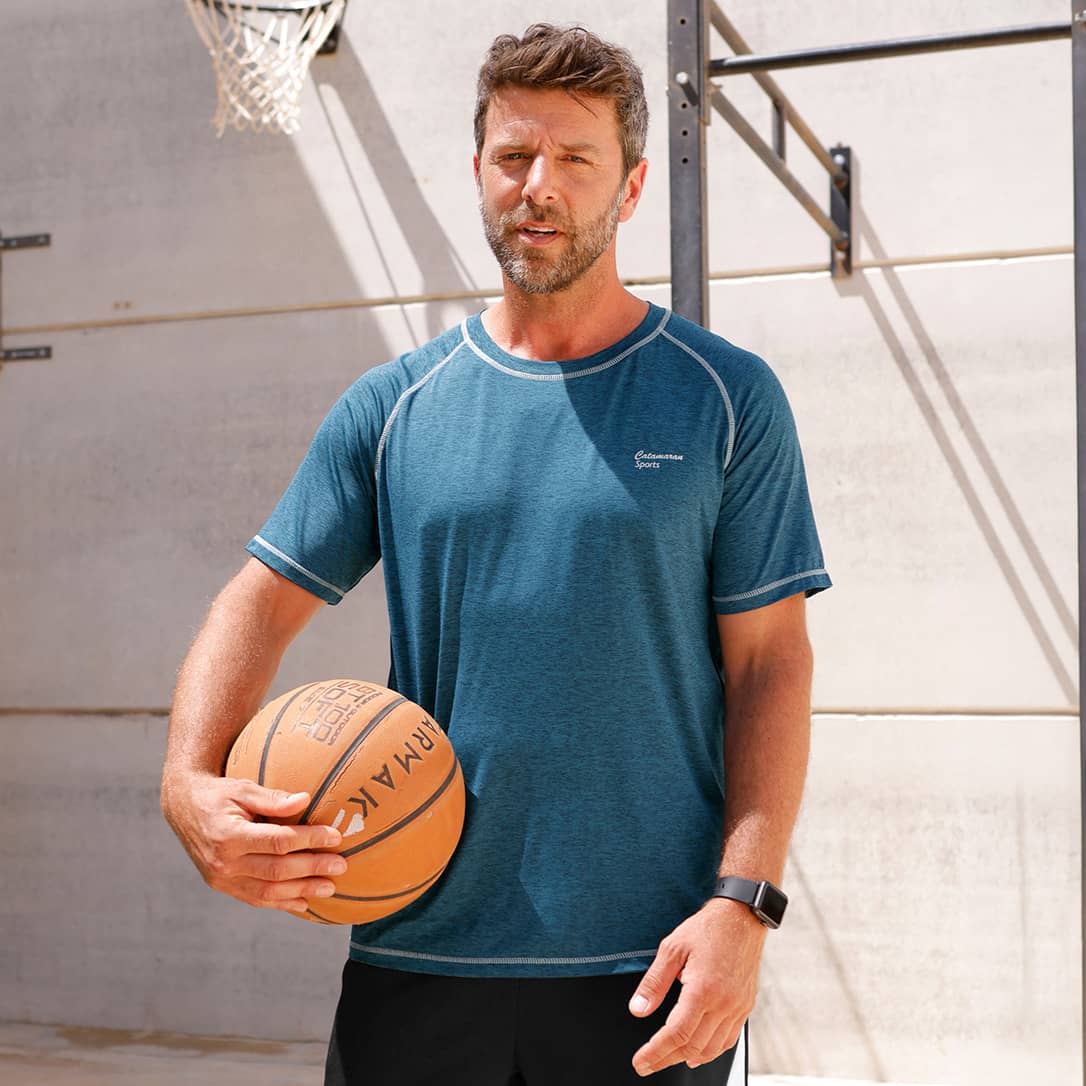 Ein Mann mit kurzem Haar und Bart steht auf einem Basketballplatz, trägt ein blaues Sportshirt und hält einen Basketball.