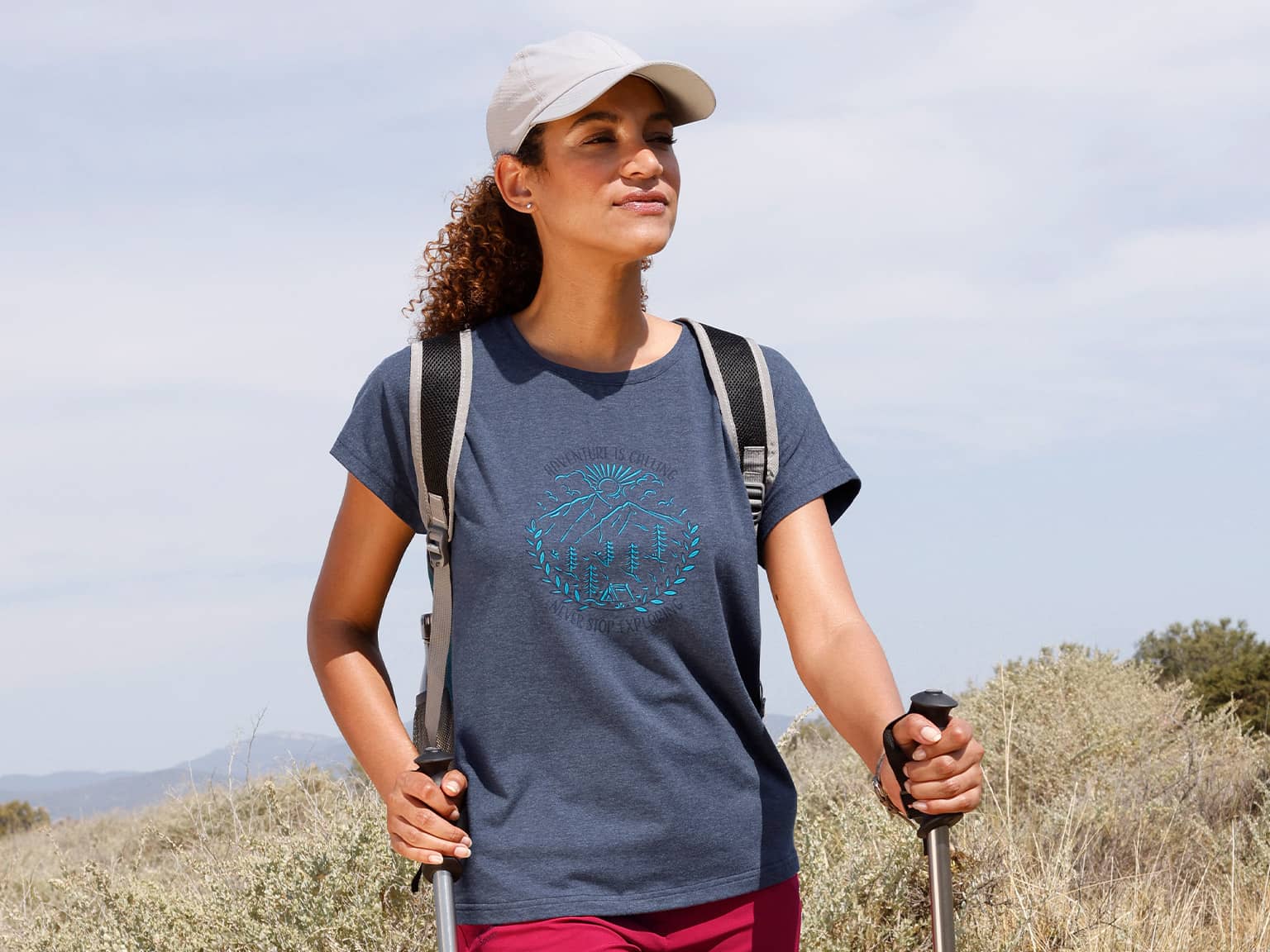 Ein Model mit lockigem Haar trägt ein dunkelblaues T-Shirt, eine weiße Kappe und einen Rucksack, hält Wanderstöcke und steht in einer offenen Landschaft.