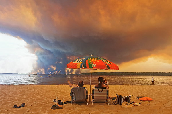 Beach goers resting on a beach look out across the water as clouds of smoke rise from the Gironde wildfire on the opposite shore. 