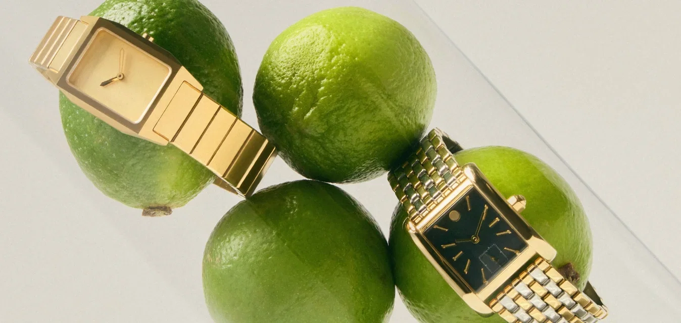 Seis limas verdes dentro de un cilindro de vidrio transparente con dos relojes de pulsera dorados descansando sobre las limas.