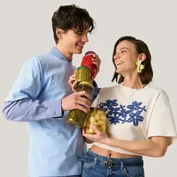 Giovane uomo e donna sorridenti, che tengono barattoli di verdure sottaceto, in piedi contro uno sfondo chiaro e semplice.