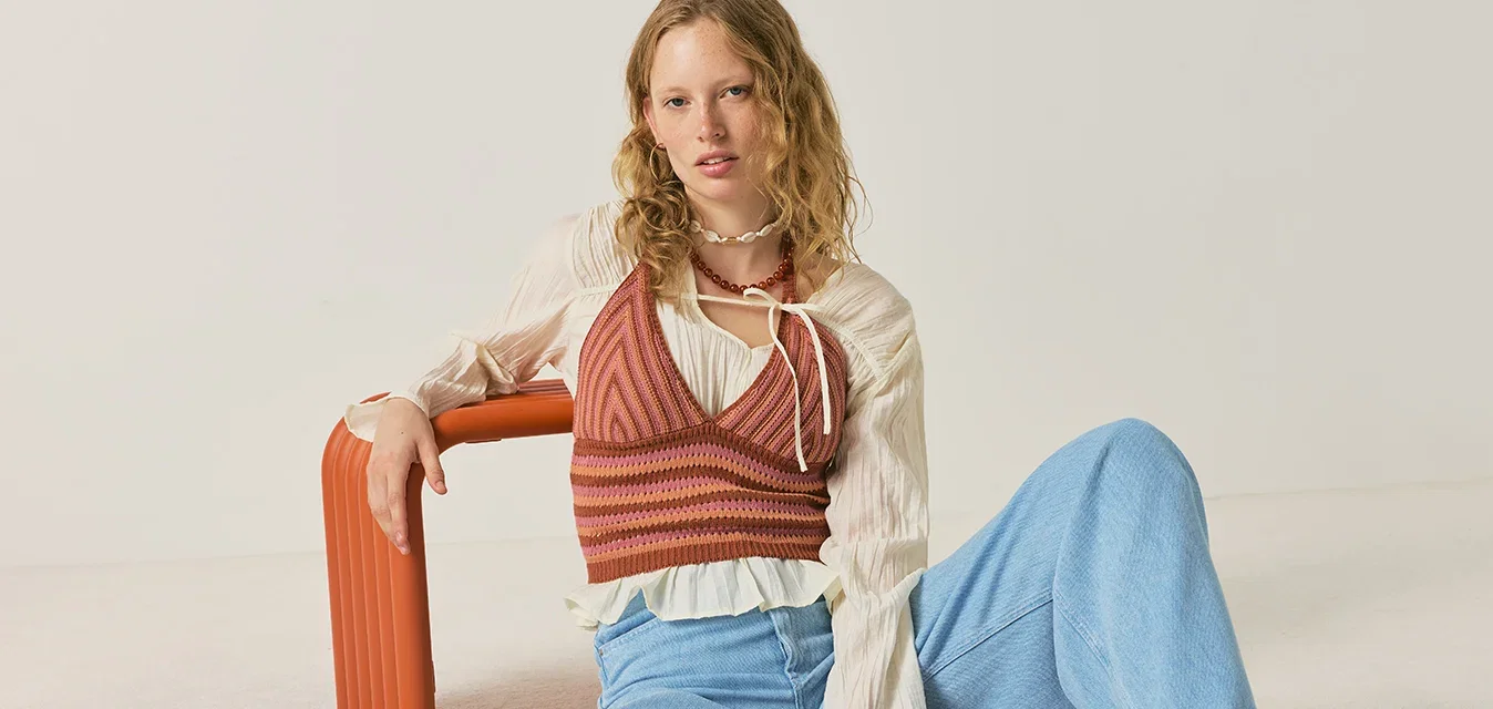 Giovane donna con capelli mossi seduta sul pavimento che indossa un gilet a righe, camicetta bianca, pantaloni larghi blu e scarpe intrecciate marroni, appoggiata a una panchina arancione.