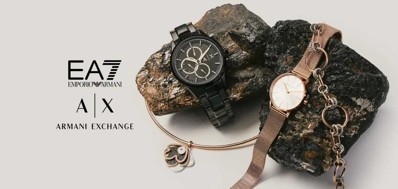 Due orologi da polso, uno in metallo nero e uno in rete color oro rosa, posati su rocce scure e strutturate su uno sfondo chiaro.