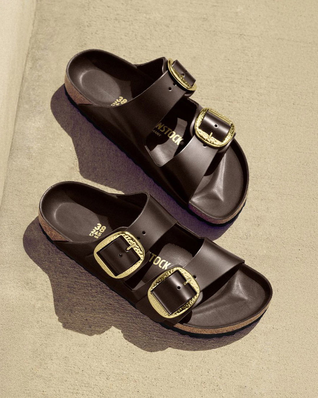 Pair of black double-strap cork-soled sandals with large gold buckles displayed on textured concrete