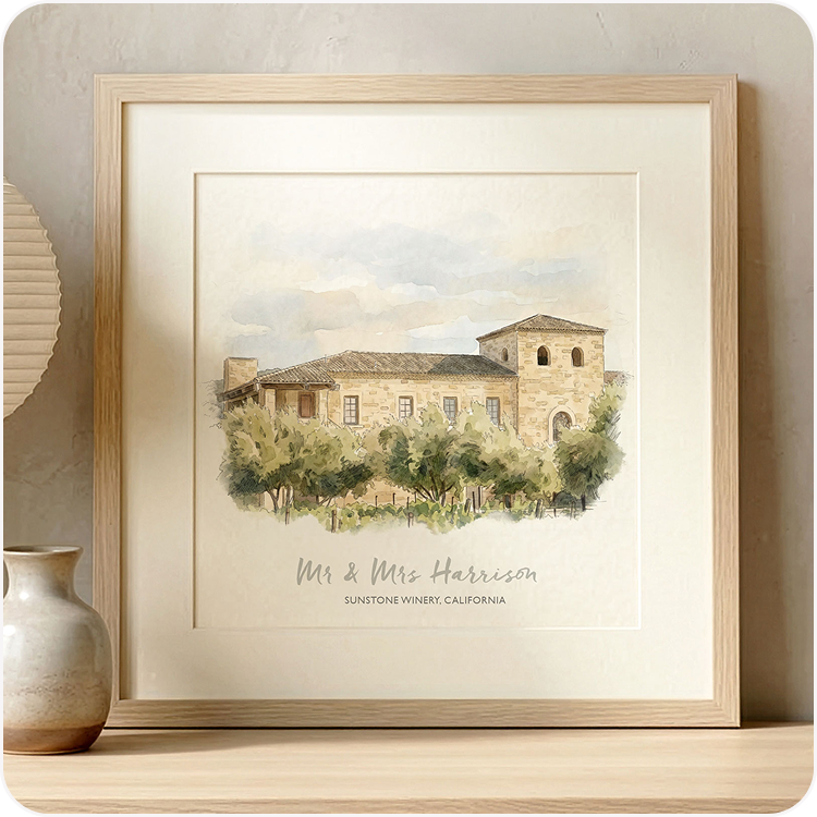 A framed watercolour illustration of a wedding venue personalised with the couple's names, displayed on a wooden surface