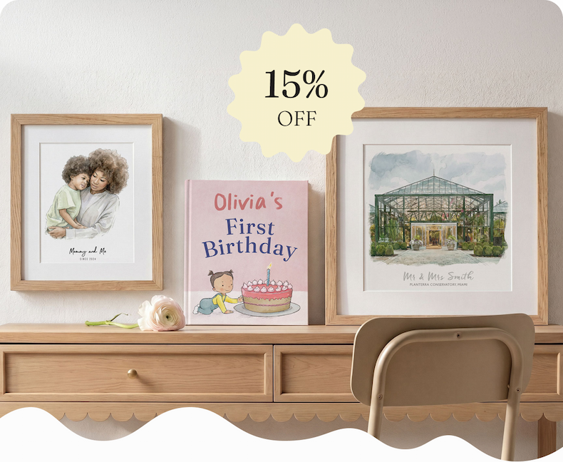 A framed watercolour illustration of a mother and child, a personalised first birthday story book and a framed watercolour wedding venue sketch, displayed on a wooden sideboard with a flower, with a 15% off badge overlay
