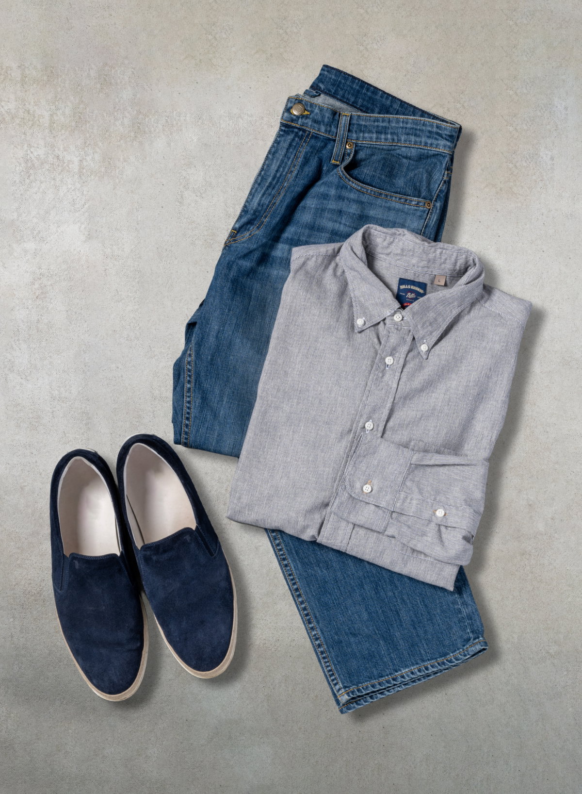A Bills Khakis standard fit shirt and Bills Khakis denim pant sit nextto casual blue slip on sneakers on a light beige grey surface