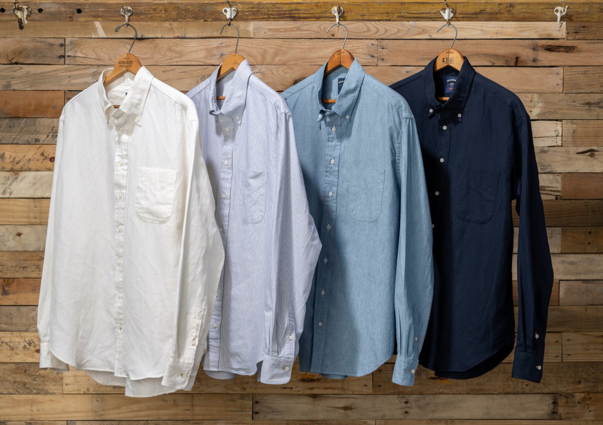 Four colors of Bills Khakis tandard fit shirts hang against a wooden plank backdrop