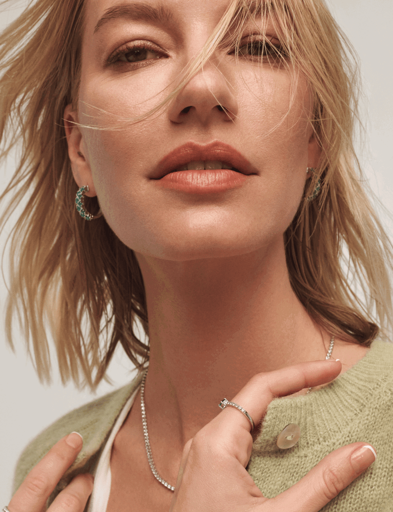 Woman in light green cardigan, diamond tennis necklace, green gemstone hoop earrings and ring.